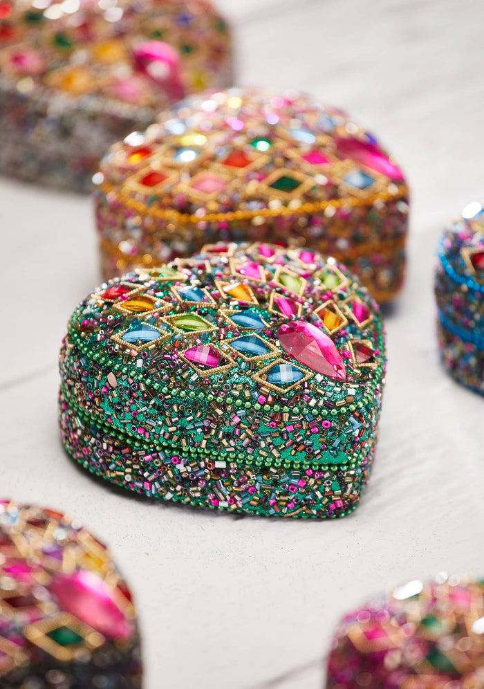 A close-up of a green beaded and rhinestone-embellished heart-shaped decorative piece, with other similar pieces blurred in the background on a light surface.