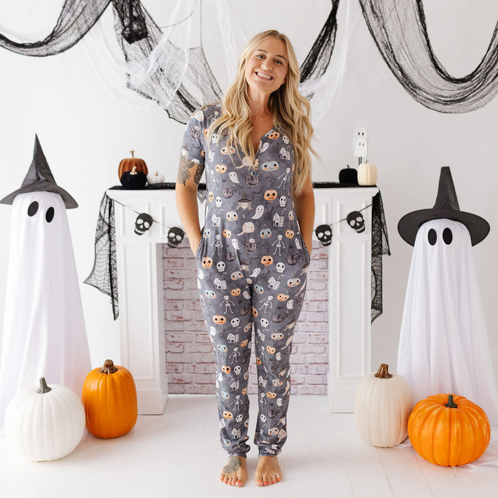 A woman stands barefoot in a Halloween-themed room wearing a gray short-sleeve pajama set with an all-over ghost and owl print. The room is decorated with black spiderwebs, white ghost decorations, pumpkins, and a white brick fireplace.