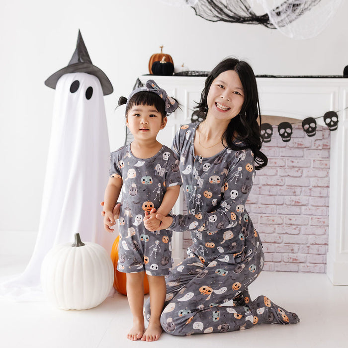 A woman and a young child wearing matching gray Halloween pajamas with a ghost and pumpkin print, kneeling in a decorated room with a white fireplace, spiderweb decorations, and pumpkins.