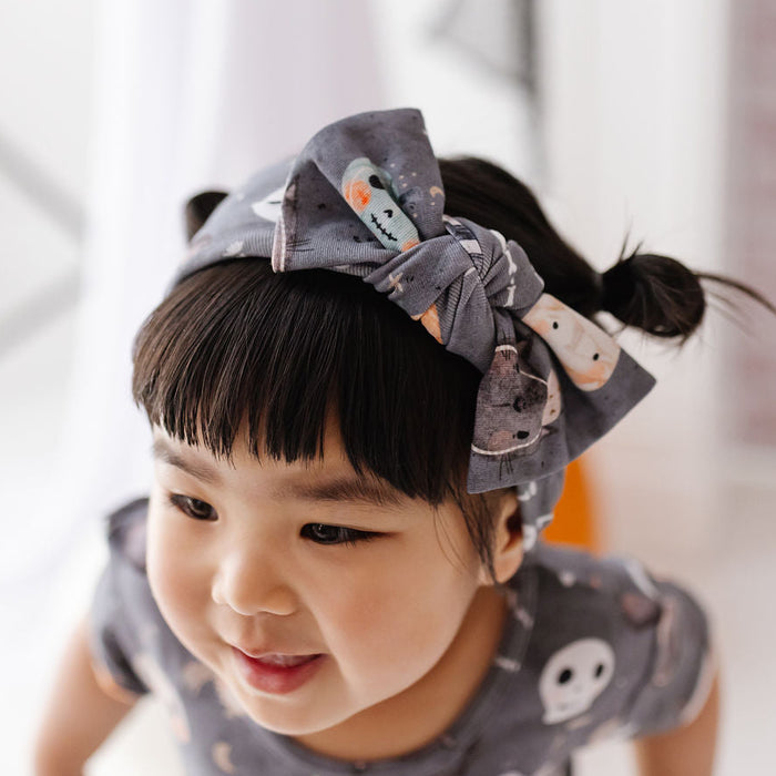 A young child wearing a gray fabric bow headband with cartoon animal prints and a matching gray top, photographed in a bright indoor setting.