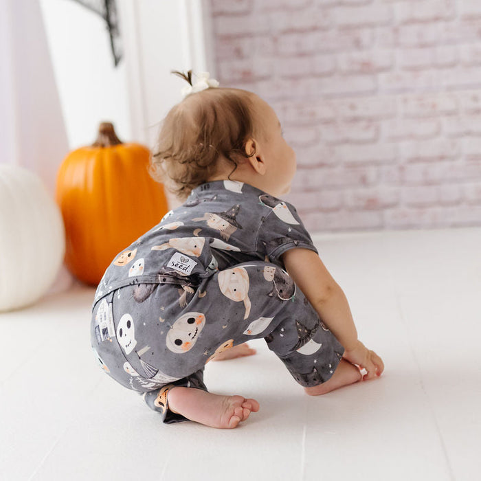 A baby wearing a gray short-sleeve romper with an owl pattern, crouching on a white wooden floor in a styled room with pumpkins and a brick wall.