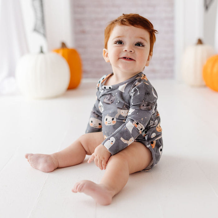 A baby sitting on a white floor wearing a gray long-sleeve romper with a cute ghost and owl pattern, surrounded by Halloween decorations like pumpkins and ghost props.
