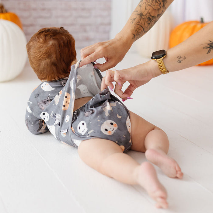 A baby wearing a gray reusable cloth diaper with a white and orange ghost pattern lies on a white floor while an adult's hands adjust the diaper. Pumpkins and a brick wall are visible in the background.