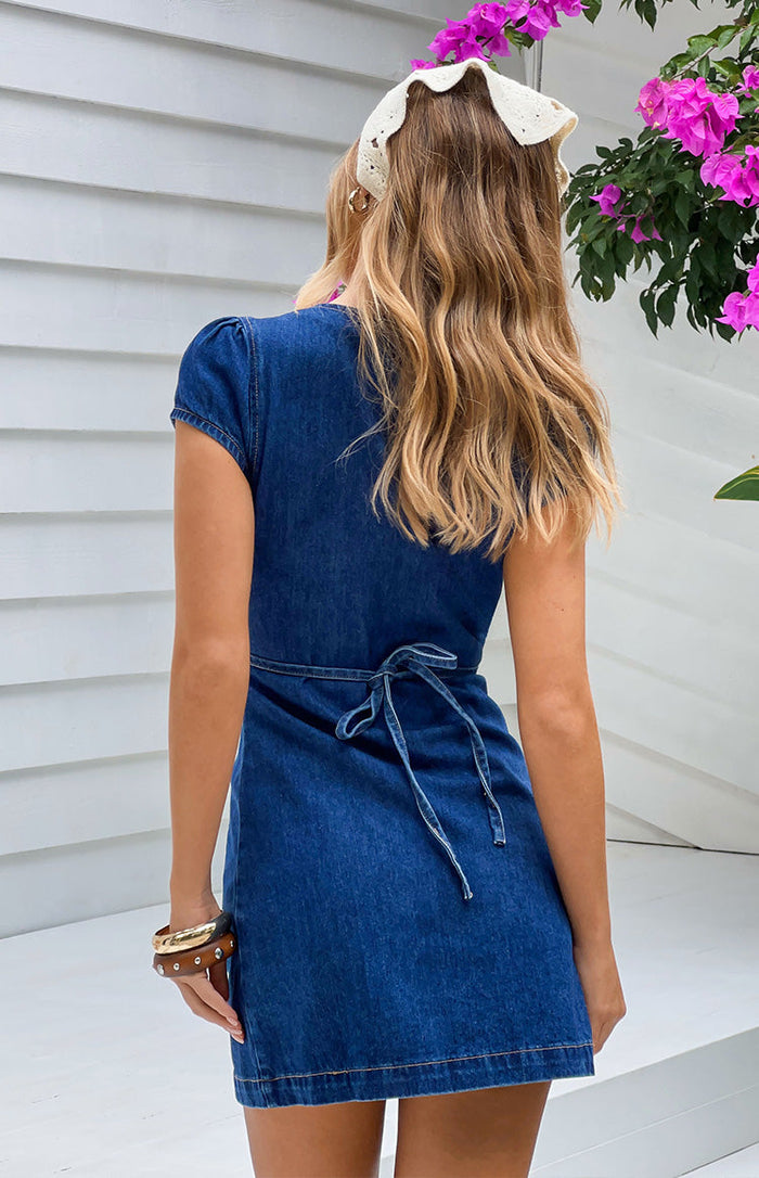 Rear view of a dark blue denim mini dress with short puff sleeves and a tie-back waist detail