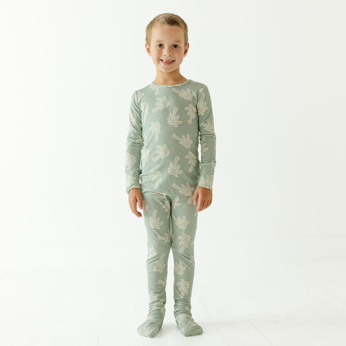 A young child standing against a plain white studio background, wearing a matching long-sleeve top and leggings pajama set in a pale sage green color with a light beige palm tree pattern.