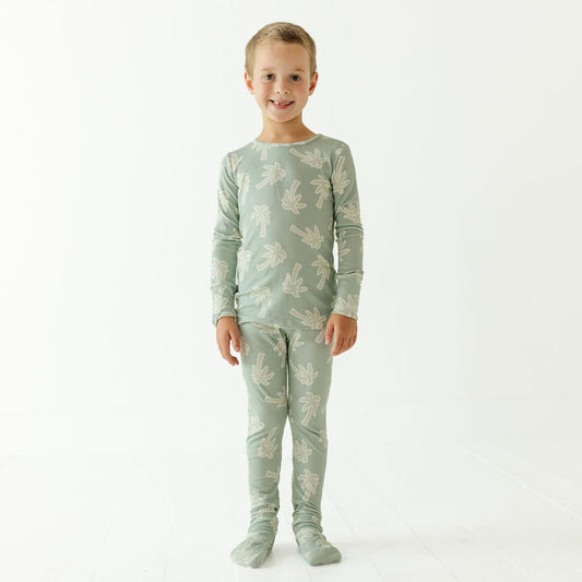 A young child standing against a plain white studio background, wearing a matching long-sleeve top and leggings pajama set in a pale sage green color with a light beige palm tree pattern.