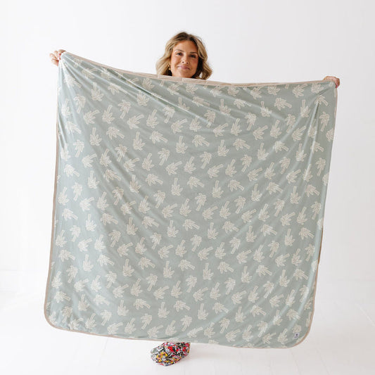 A woman holds up a large square blanket or throw with a sage green background and a repeating white leaf pattern, shown against a plain white studio background.