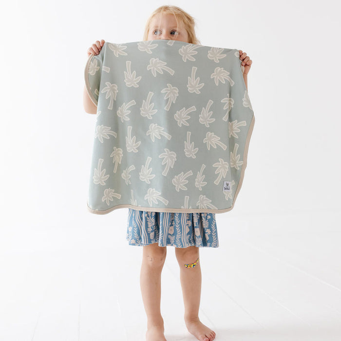 A young child stands barefoot against a white studio background, holding up a square cloth or blanket with a pale sage green base and a repeating cream palm tree pattern. The child is wearing a blue patterned dress.