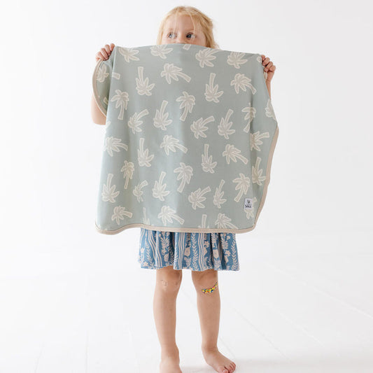 A young child stands barefoot against a white studio background, holding up a square cloth or blanket with a pale sage green base and a repeating cream palm tree pattern. The child is wearing a blue patterned dress.