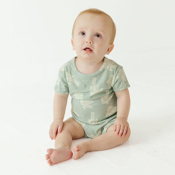 A baby sitting on a white studio floor wearing a sage green short-sleeve bodysuit with a light beige leaf pattern.