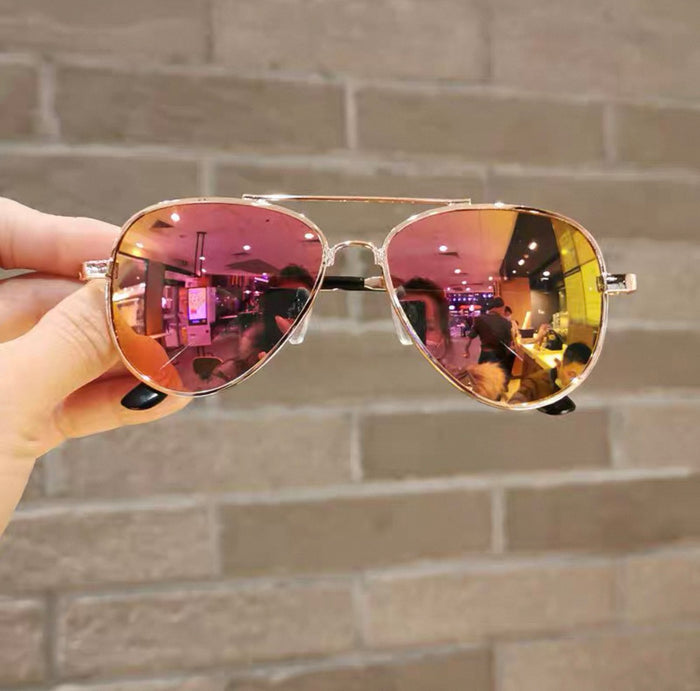 Aviator sunglasses with pink and gold reflective lenses held against a brick wall.