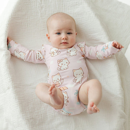 A baby lying on a textured white muslin blanket, wearing a light pink long-sleeve bodysuit with a cute cartoon cat print.