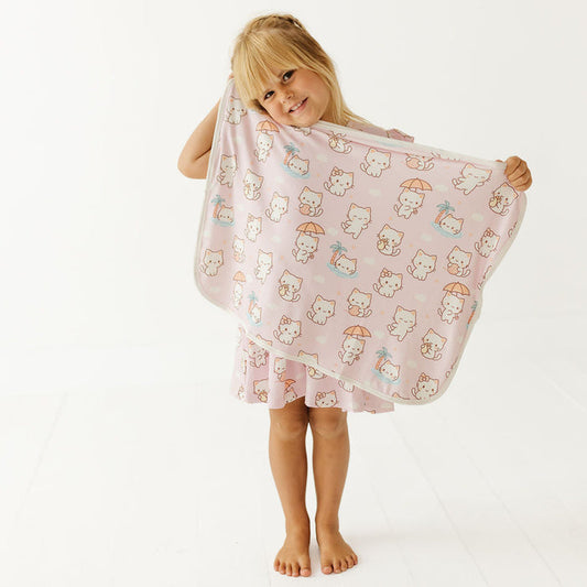A young girl with blonde hair stands barefoot against a white studio background, holding up a light pink blanket or towel patterned with cute cartoon cats in white, orange, and blue.