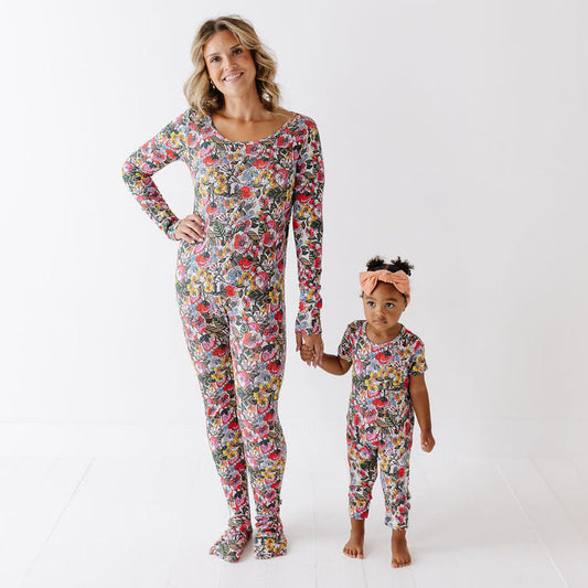 A woman and a young child standing hand in hand, both wearing matching long-sleeve pajama sets with a dense multicolor floral print, against a clean white studio background.