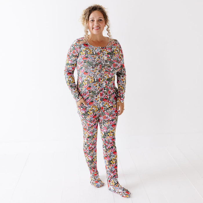 A woman modeling a long-sleeve footed pajama set with a dense multicolor floral and abstract print, standing against a plain white studio background.
