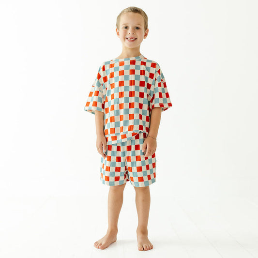 A young boy stands barefoot against a plain white studio background, wearing a matching short-sleeve pajama set with an orange, red, and light blue checkerboard pattern.