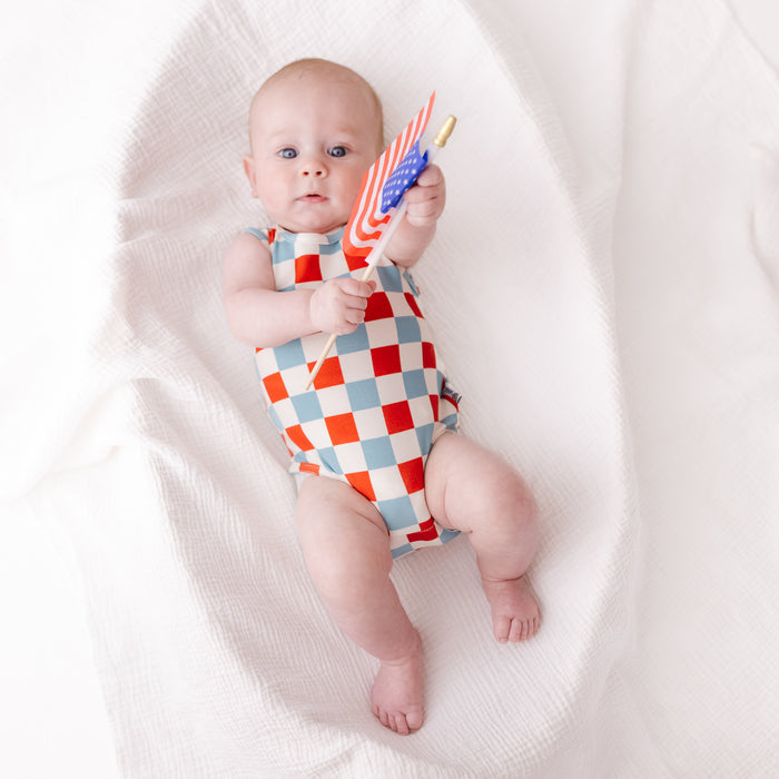 A baby lying on a white textured blanket, wearing a red, white, and light blue checkered sleeveless bodysuit and holding a small toy flag.