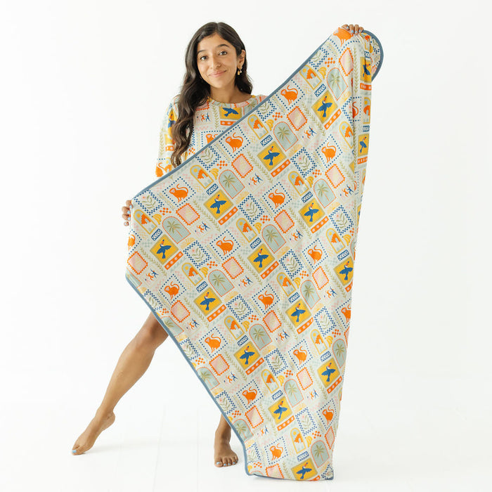 A woman stands barefoot against a white studio background, holding up a large patterned blanket or throw. The blanket has a cream base with repeated geometric and abstract motifs in orange, blue, and muted green, plus a blue border.