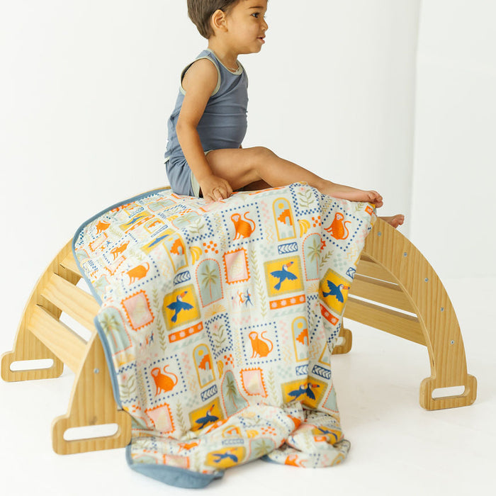 A young child sits on a light wood climbing arch while a patterned blanket is draped over the structure. The child is wearing a blue-gray tank top, and the blanket has a white base with blue, orange, and yellow dinosaur and square motifs.