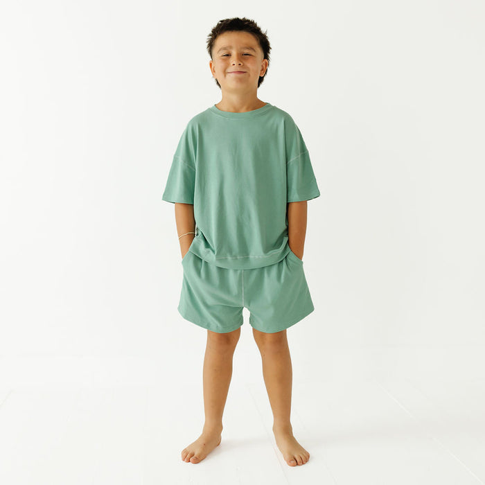 A young boy stands barefoot against a plain white studio background, wearing a matching sage green lounge set consisting of a short-sleeve t-shirt and shorts.