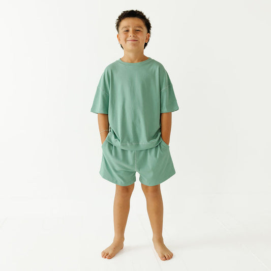 A young boy stands barefoot against a plain white studio background, wearing a matching sage green lounge set consisting of a short-sleeve t-shirt and shorts.
