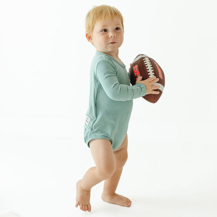 A toddler standing barefoot on a white studio background, wearing a long-sleeve sage green romper and holding a brown American football.