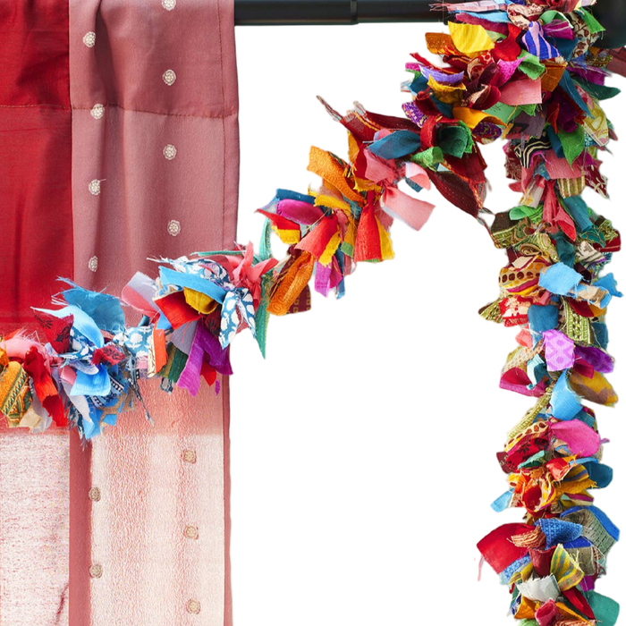 A close-up of a colorful decorative garland made of many layered fabric strips in bright shades, draped along the top and right side of a frame next to a pink fabric panel.