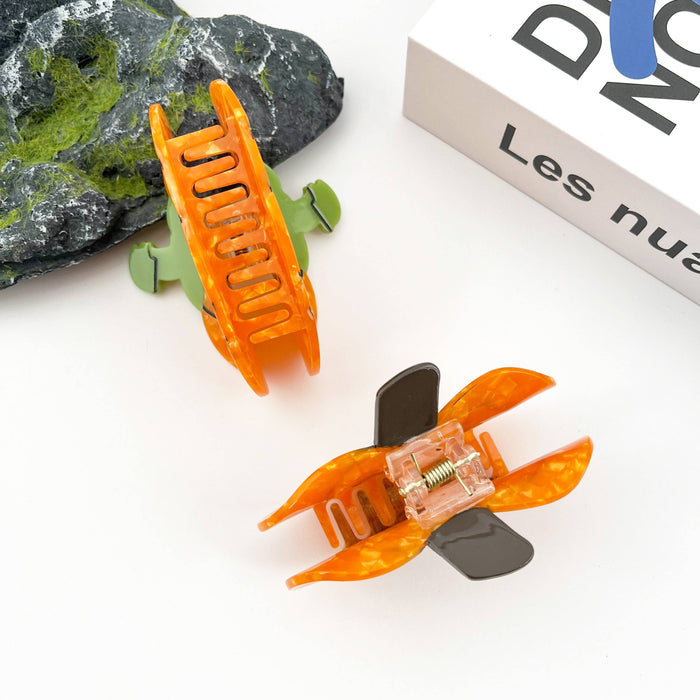 Two hair claw clips styled on a white surface with a dark green rock and a white box in the background. One clip is translucent orange with a dark gray wing-like accent, and the other is translucent green with a light green wing-like accent.