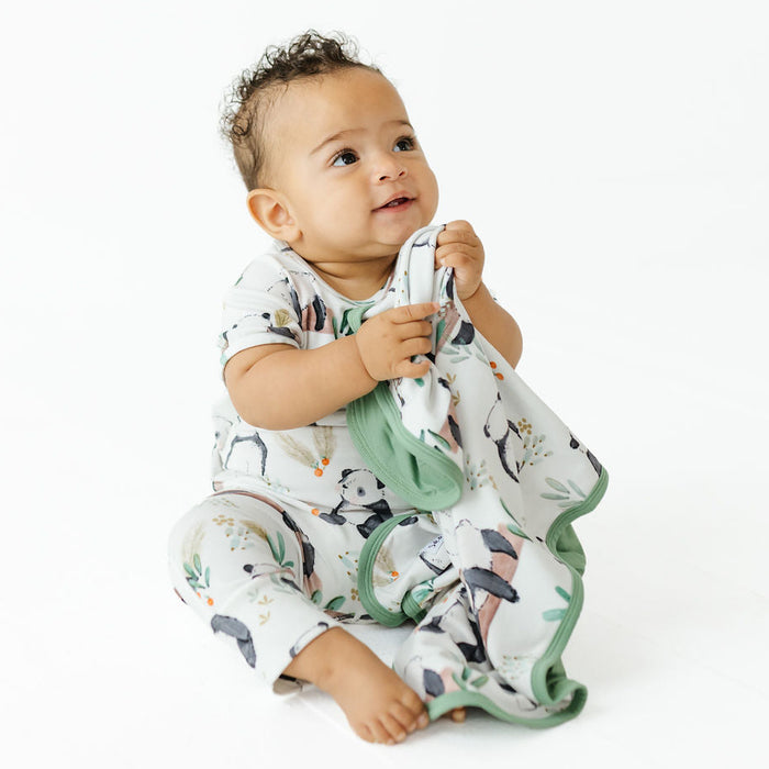 A baby sitting on a white studio background, wearing a white short-sleeve romper and matching blanket set with a penguin print and sage green trim.
