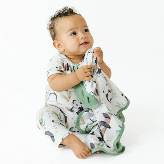A baby sitting on a white studio background, wearing a white short-sleeve romper and matching blanket set with a penguin print and sage green trim.