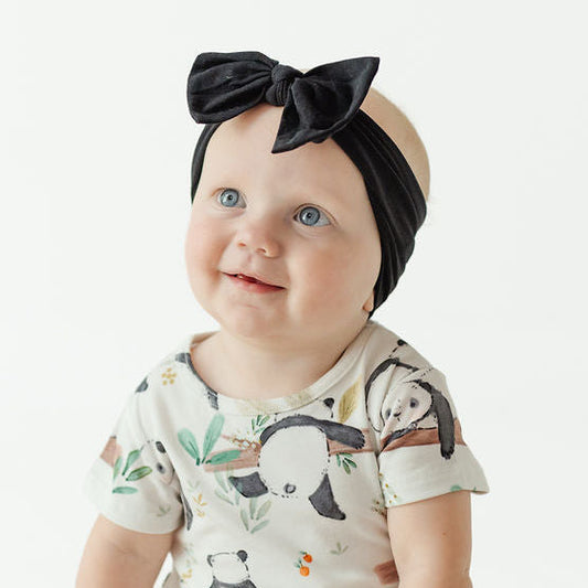A baby sitting on a white studio floor wearing a short-sleeve cream bodysuit with a panda print and a black velvet bow headband.