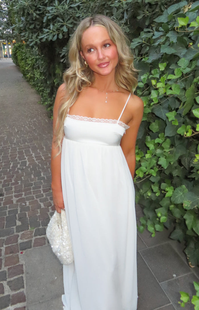 White satin maxi dress with a straight neckline, delicate lace trim, thin spaghetti straps, and empire waist