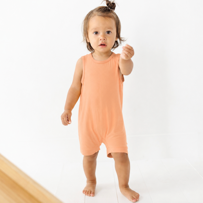 A toddler girl wearing a peach sleeveless romper jumpsuit, standing barefoot against a clean white studio background.