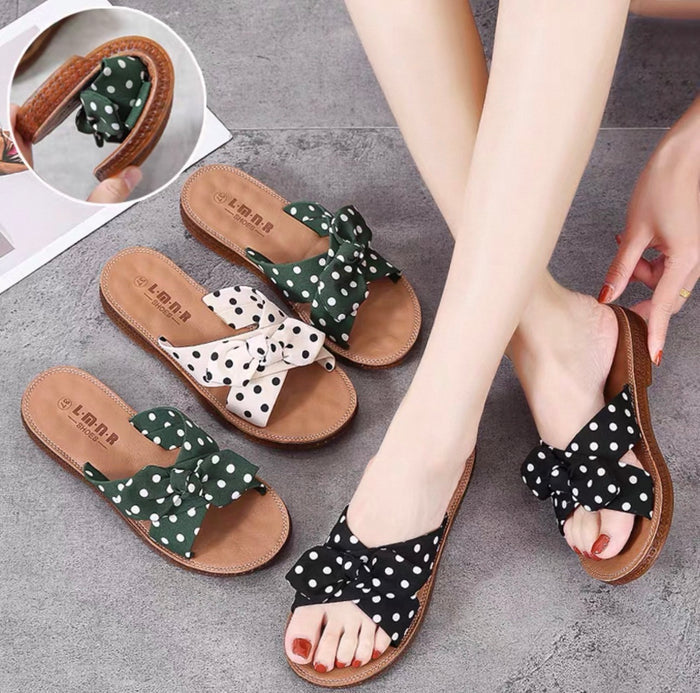 Pair of polka dot sandals with bows worn by a person on a gray floor.