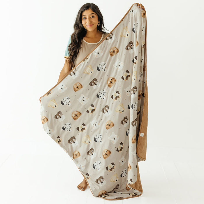 A woman holds up a large fleece blanket featuring a pattern of cartoon dog faces on a light beige background, with a solid brown reverse side, against a plain white studio background.