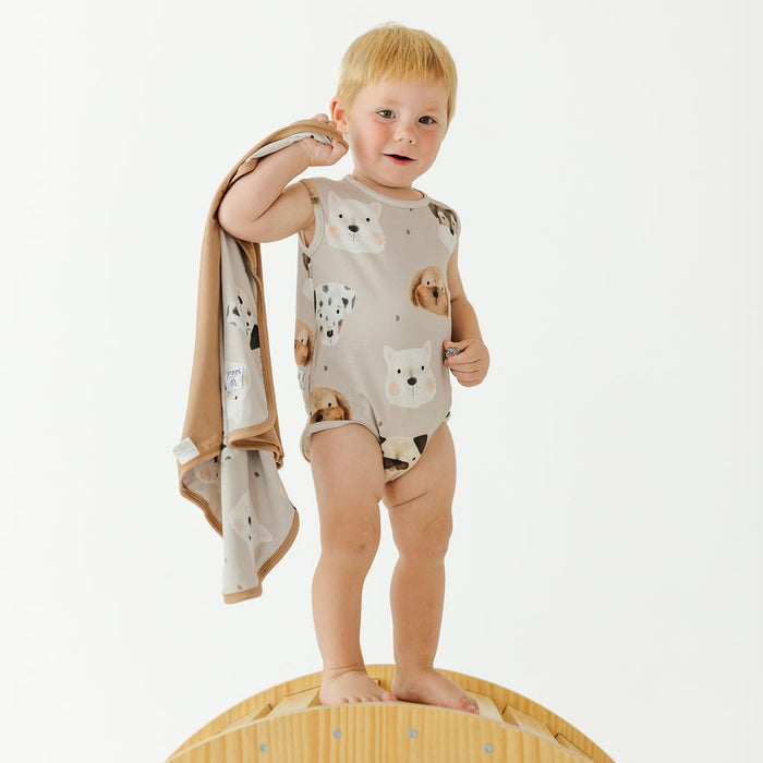 A toddler stands barefoot on a wooden arch in a studio setting, wearing a sleeveless beige romper with a dog print and holding a matching beige towel.
