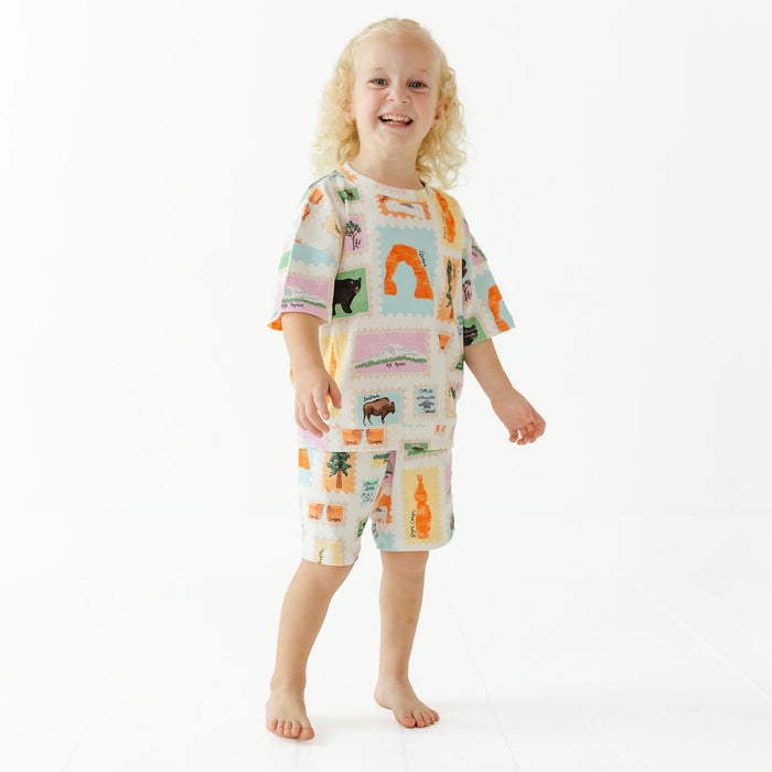 A young child with curly blonde hair stands barefoot against a plain white studio background, wearing a matching short-sleeve pajama set with a colorful patchwork print of palm trees, suns, and animals.