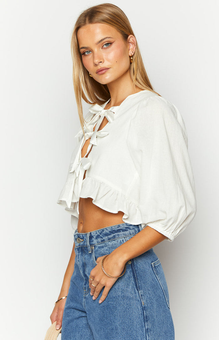 Side view of a white cropped tie up top with front bow details, puffed sleeves, and a ruffled hem
