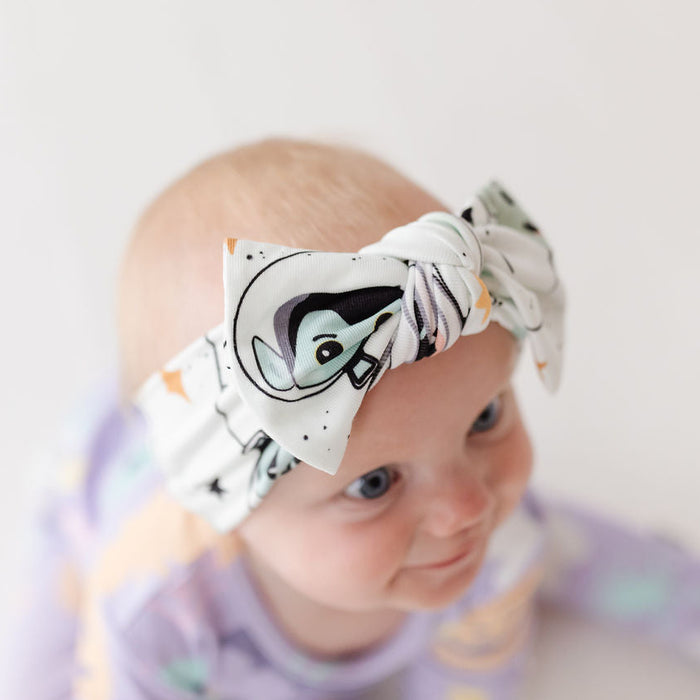 A baby wearing a knotted fabric bow headband with a penguin face print, photographed against a soft white background.