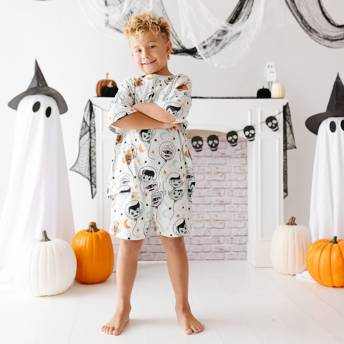 A young boy stands barefoot in a Halloween-themed room, wearing a white short-sleeve nightshirt or pajama top with a cartoon skull-and-miscellaneous print. The room has white walls, a white fireplace mantel, black spiderweb decorations, ghost cutouts, pumpkins, and black witch hats.