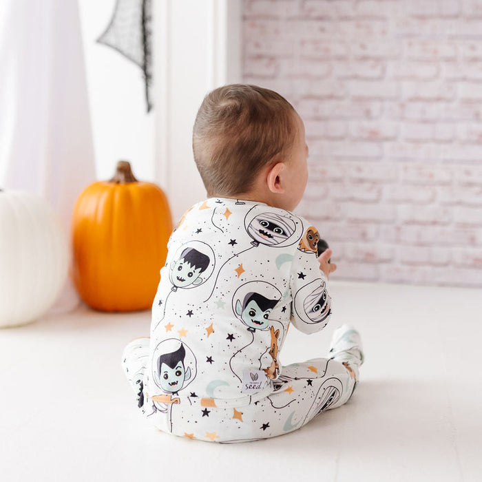 A baby sits on a white floor facing a decorated fireplace, wearing a white footed sleeper with a cartoon astronaut print. A white pumpkin, an orange pumpkin, and a ghost decoration are visible in the background.