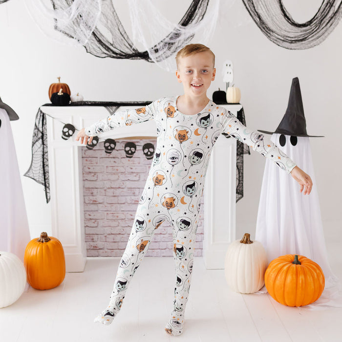 A young boy stands in a Halloween-themed room wearing a white long-sleeve footed pajama with a black, orange, and gray cartoon cat pattern. He is posed in front of a white fireplace mantel decorated with black spiderwebs, ghost figures, pumpkins, and skull garlands.