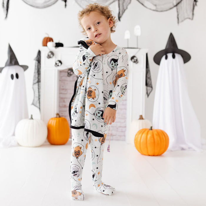 A toddler stands in a Halloween-themed room wearing a long-sleeve footed pajama set with a white base and a cartoon spider pattern. The room features a white fireplace mantel decorated with black spiderwebs, orange pumpkins, and ghost props.