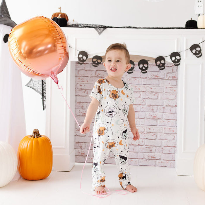 A toddler stands barefoot in a Halloween-themed room, wearing a white one-piece pajama with a cartoon animal print. The child is holding a large metallic rose-gold foil balloon, with pumpkins, black-and-white ghost figures, and a brick-patterned fireplace backdrop behind them.