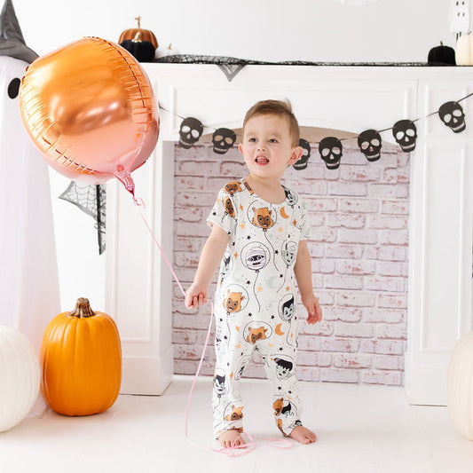 A toddler stands barefoot in a Halloween-themed room, wearing a white one-piece pajama with a cartoon animal print. The child is holding a large metallic rose-gold foil balloon, with pumpkins, black-and-white ghost figures, and a brick-patterned fireplace backdrop behind them.