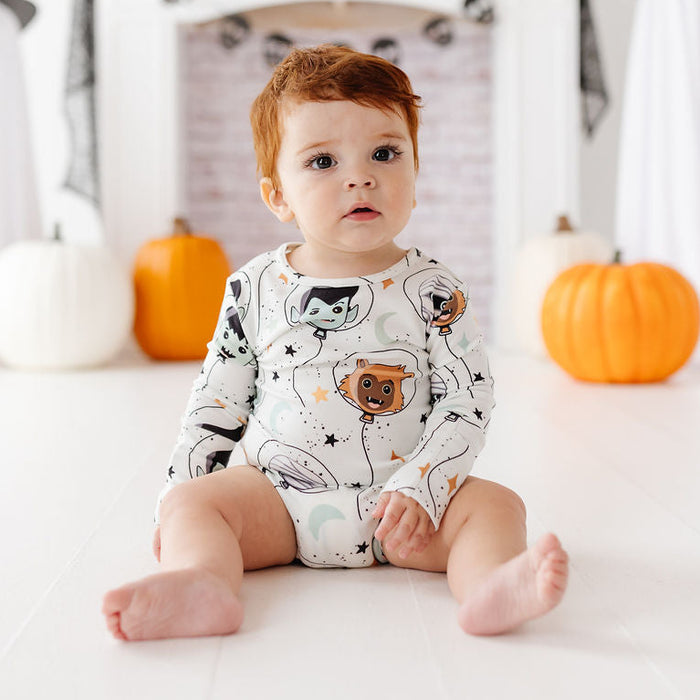 A baby sitting on a white floor in a Halloween-themed room, wearing a white long-sleeve onesie with a cartoon astronaut and cat print. Pumpkins, ghost decorations, and a white fireplace mantel are visible in the background.