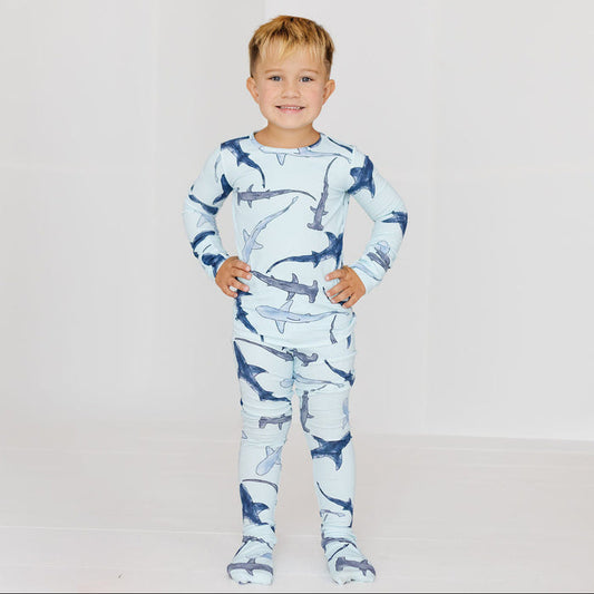 A young boy stands with hands on hips wearing a long-sleeve footed pajama set with a shark print, set against a plain white studio background.