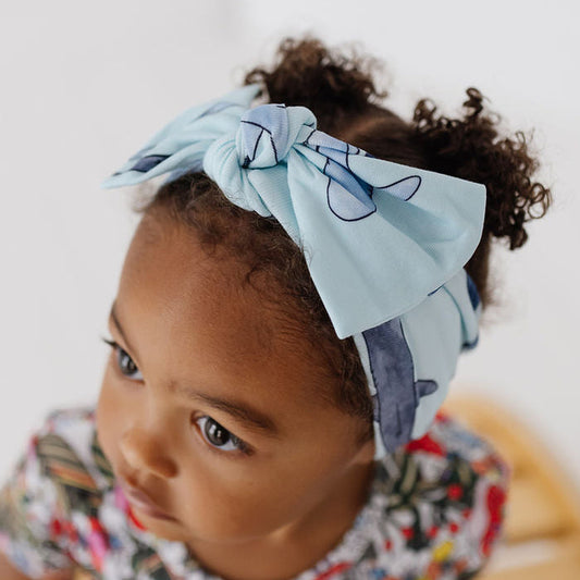 A toddler girl with curly dark hair wearing a light blue fabric bow headband with a dark blue abstract pattern, paired with a colorful floral dress, sitting on a wooden slatted bench against a white wall.