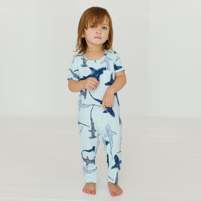 A toddler stands barefoot in a bright white studio setting, wearing a short-sleeve one-piece pajama or lounge set with an allover shark print on a pale blue background.