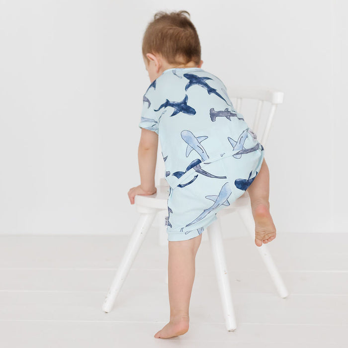 A baby wearing a light blue short-sleeve romper with a shark print, seen from behind as they climb a white wooden chair in a bright, minimalist room.